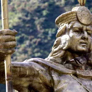 A close-up of a stone statue of an Inca figure holding a staff, with doves perched on its shoulder and a lush green mountain in the background, representing the rich history encountered on a Machu Picchu Express trip. - Qosqo Expeditions